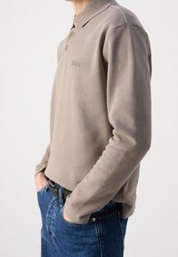 Person wearing beige long-sleeve polo with "BOSS" logo and blue jeans, hands in pockets, against a plain white background.