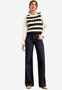 Striped knit sweater in black and cream, layered over a white blouse with ruffled sleeves. Paired with dark blue flared jeans and beige shoes.