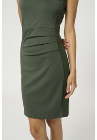 Robe verte ajustée avec un panneau latéral côtelé, texture lisse et plis subtils à la taille. La longueur tombe au-dessus du genou.