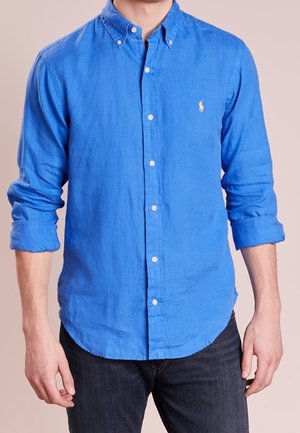 Man wearing a bright blue, long-sleeve button-up shirt with sleeves rolled up, paired with dark jeans, against a plain background.