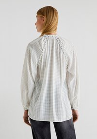Blusa bianca con texture, maniche lunghe, con dettagli arricciati sulla schiena e un design a motivi sottili.