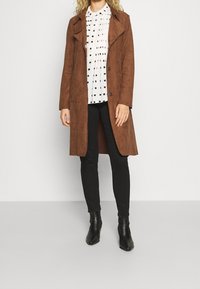 Manteau en daim marron avec col à revers, fermeture à boutons noirs, associé à un chemisier blanc à pois noirs, un pantalon slim noir et des bottines.