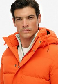Jasnopomarańczowa ocieplana kurtka z kapturem, z zamkiem błyskawicznym, napami oraz teksturowaną białą warstwą wewnętrzną. Gładki, wytrzymały materiał.
