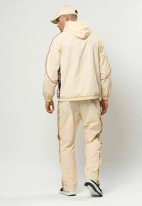 Beige vindjakke med røde detaljer og elastiske manchetter, kombineret med matchende bukser med sidestriber, båret med en beige kasket.