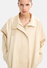 Beżowa, oversize'owa kurtka z szerokim kołnierzem, zamek błyskawiczny oraz kieszeniami z przodu; wykonana z gładkiego, lekkiego materiału o miękkiej teksturze.