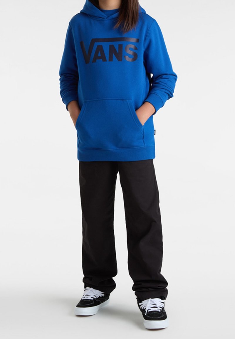 Personne portant un sweat à capuche bleu Vans avec les mains dans la poche kangourou, pantalon noir et baskets noires et blanches, debout sur un fond blanc.