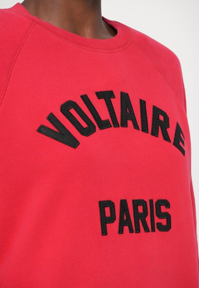 Rood sweatshirt met zwarte tekst die "VOLTAIRE" boven "PARIJS" leest, met een ronde hals en raglanmouwen. Zachte stoftextuur.
