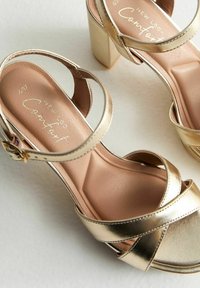 Sandalias de tacón alto metálicas doradas con correas cruzadas, un tacón grueso y una plantilla suave de color beige que presenta el nombre de la marca en una tipografía decorativa.