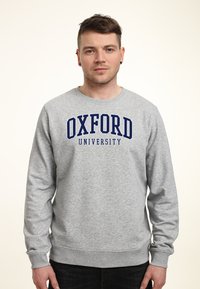 Grå sweatshirt med långa ärmar, rund halsringning, har "OXFORD UNIVERSITY" i fetblå bokstäver över bröstet. Mjuk textur, avslappnad passform.