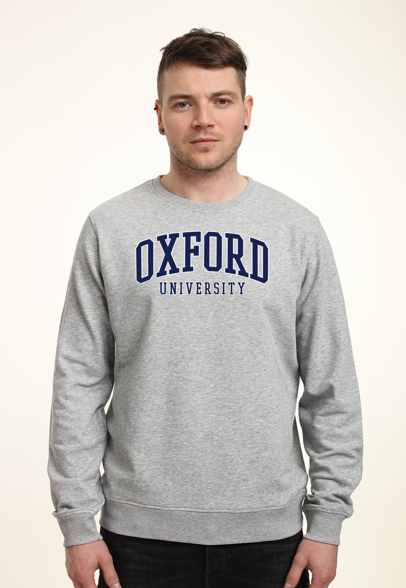 Grå sweatshirt med långa ärmar, rund halsringning, har "OXFORD UNIVERSITY" i fetblå bokstäver över bröstet. Mjuk textur, avslappnad passform.