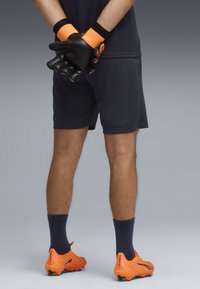 Guantes de portero negros con correas de muñeca naranjas, combinados con botas de fútbol naranjas y pantalones cortos azul marino, vistos desde atrás.