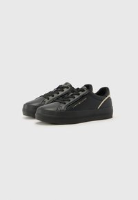 Baskets en cuir noir avec une surface texturée, dotées de lacets noirs, d'une semelle plate et d'un accent de logo doré sur le côté. Design simple.