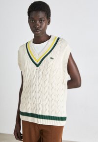 Gilet en tricot torsadé crème avec un col en V, présentant des rayures vertes et jaunes le long du col et une bande verte à l'ourlet.