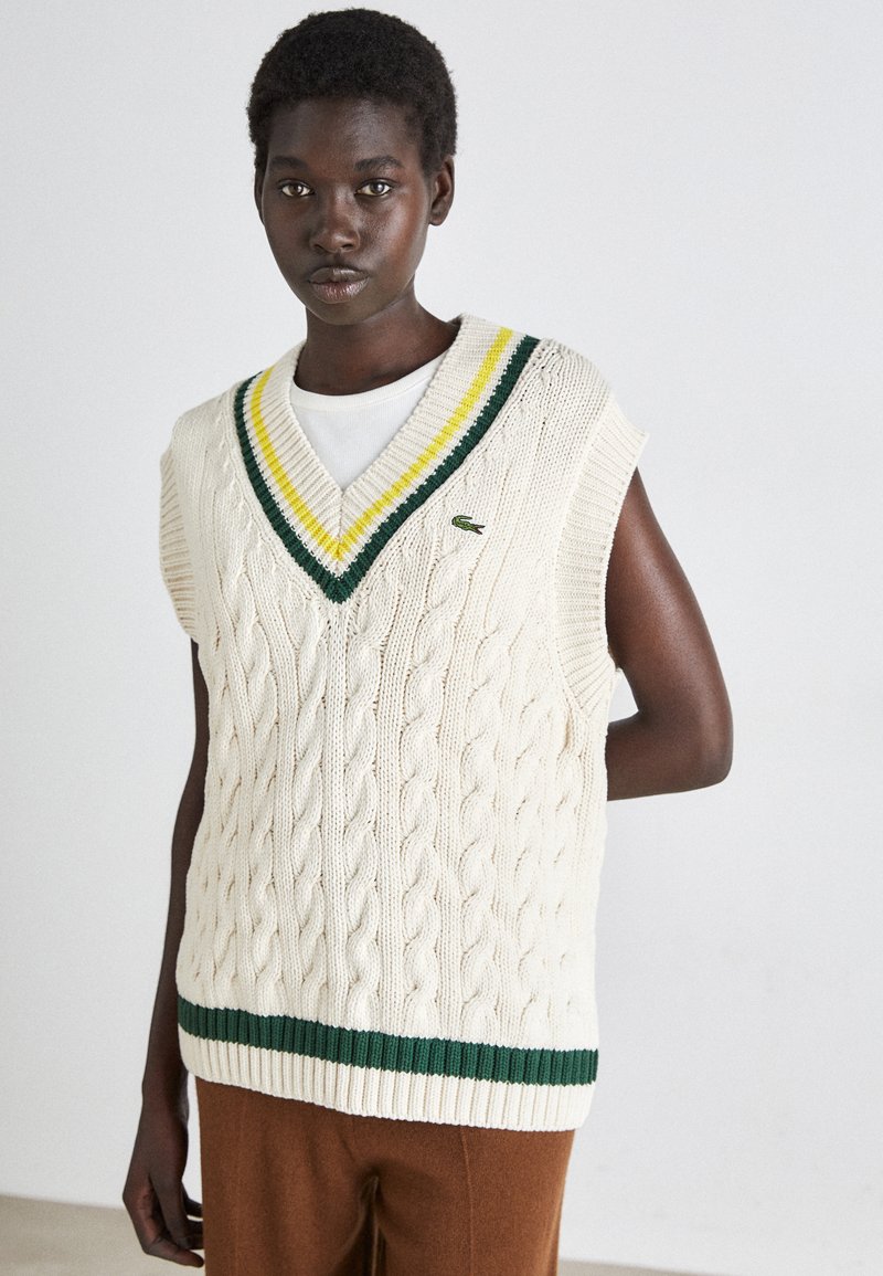 Gilet en tricot torsadé crème avec un col en V, présentant des rayures vertes et jaunes le long du col et une bande verte à l'ourlet.