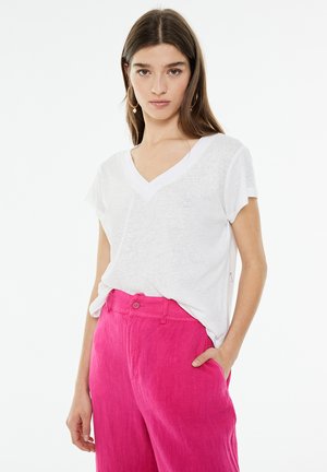 T-shirt bianco con scollo a V realizzato in tessuto leggero e leggermente trasparente, abbinato a pantaloni di lino rosa brillante con chiusura a bottone.