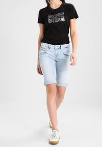 T-shirt noire avec un motif en sequins, shorts en denim bleu clair avec des ourlets retroussés, et baskets blanches avec des semelles beiges.