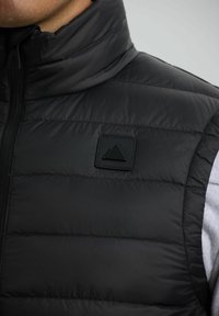 Close-up van een zwarte gewatteerde vest met hoge kraag en vierkant logo-patch met twee overlappende driehoeken op de borst.