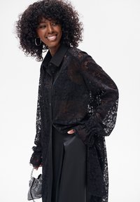 Czarna bluzka z długim rękawem z koronki z zapinaniem na guziki. Posiada kołnierzyk, luźny fason i misterny wzór koronki. Połączona z czarnymi spodniami.