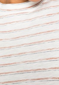 Lähellä oleva kuva valkoisesta kankaasta, jossa on ruosteisia ja mustia vaakasuoria raitoja, henkilön rintakehässä ja kaula-alueella.