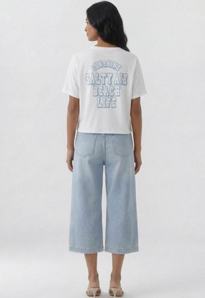 Frau mit langen schwarzen Haaren, die ein weißes, kurz geschnittenes T-Shirt mit Strandmotiv-Text und hellblaue, weit geschnittene Jeans trägt, steht mit dem Rücken zum Betrachter.