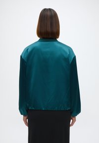 Teal selyem bomber dzseki laza szabással, leengedett vállakkal és rugalmas mandzsettákkal és alsó szegéllyel. Simaságával, gallér nélküli dizájnjával tűnik ki.