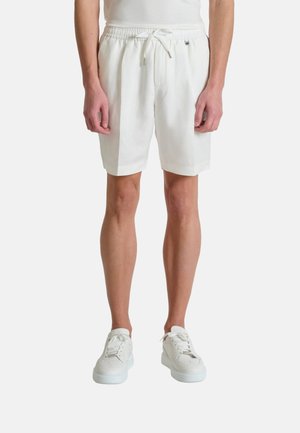 Homme portant un short blanc taille élastique avec cordon de serrage et des baskets blanches, debout devant un fond blanc uni.