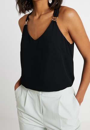 Kvinde iført en ærmeløs sort V-hals top med ringdetaljer på stropperne og lyse højtaljede bukser med hænderne i lommerne.