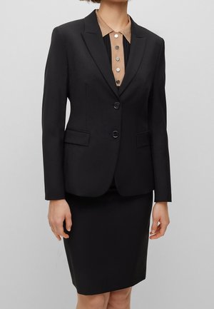 Femme portant un blazer noir à deux boutons sur une jupe noire et un polo beige avec quatre boutons métalliques.