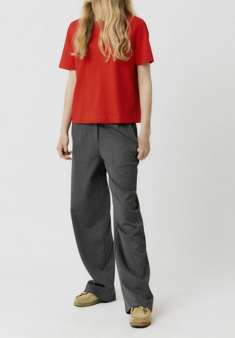 Haut rouge à manches courtes associé à un pantalon large à rayures grises. Modèle portant des chaussures plates beiges. Tissu lisse, coupe décontractée.