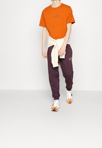 Oranžové tričko s logem, krémový svetr uvázaný v pase, tmavě fialové tepláky a pestré tenisky. Neformální sportovní oblečení.