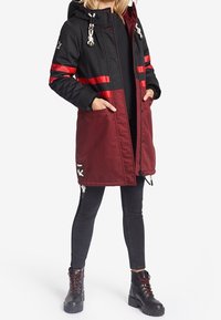 Zwarte en bordeauxrode parka met rode strepen, ritssluiting, capuchon en trekkoord. Heeft twee zijzakken en decoratieve hardware.