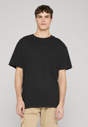 Young man stands facing forward wearing a plain black oversized t-shirt and beige cargo pants against a light gray background.