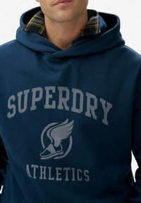 Marineblauwe sweatshirt met capuchon, met een opvallend lichtgrijs "Superdry Athletics" logo en vleugelgrafiek aan de voorkant. Textuurstof met geruite voering.