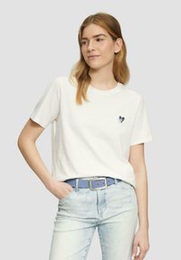 Weißes Baumwoll-T-Shirt mit kurzen Ärmeln, das ein kleines Herz-Grafik in Blau-Tönen auf der linken Brust zeigt. Getragen mit hellblauen Jeans und Gürtel.