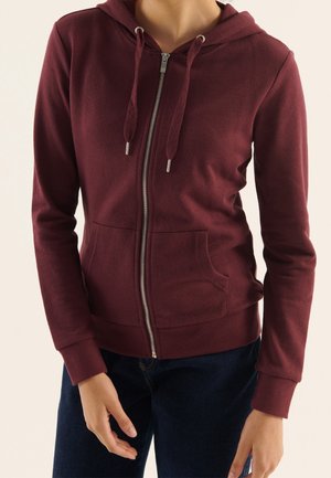 Persoon die een bordeauxrode hoodie draagt met ritssluiting, trekkoorden en zakken aan de voorkant, gecombineerd met donkerblauwe jeans.