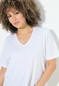 T-shirt bianco in cotone con scollo a V e maniche corte, che mostra una texture liscia. Il modello è posizionato su uno sfondo chiaro.
