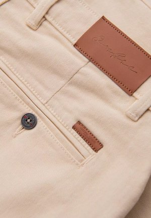 Beige denimové kalhoty s koženkovou nášivkou označenou "Signature." Mají stříbrný knoflík a malou hnědou nášivku u zadní kapsy.