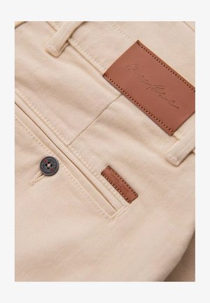 Beige denimbukser med et lædermærke mærket "Signature." Har en sølvknap og et lille brunt mærke nær baglommen.