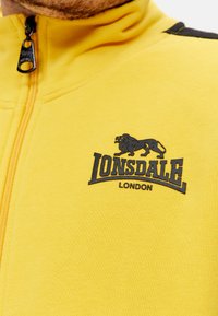 Žltá zipovacia bunda vyrobená z mäkkej tkaniny, na hrudi má vyšité čierne logo leva a text "Lonsdale London".