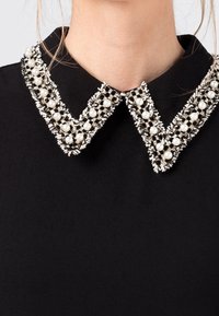 Svart plagg med en detaljerad krage prydd med vita pärlor och silverdetaljer, som visar en texturerad design med en skarp form.