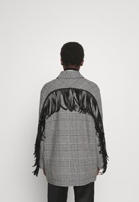 Abrigo de pata de gallo en blanco y negro con detalle de flecos negros largos a lo largo de la parte superior de la espalda, con cuello clásico y corte entallado.