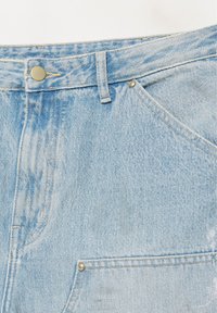 Zbliżenie jasnoniebieskich dżinsów z denimu pokazujące pas biodrowy, szlufkę na pasek, guzik, przednią kieszeń oraz nitowanie z widoczną wyblakłą fakturą.