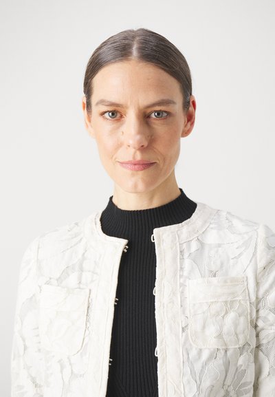 Biela čipkovaná bunda s kvetinovými vzormi, so zaobleným výstrihom, cvokovými zapínaniami a vreckom na prsníku, vrstvená nad čiernym rebrovaným topom.