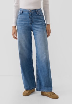 Person trägt hochtaillierte, weit geschnittene blaue Jeans mit Knopfleiste, weißes Langarmshirt und braune Mokassins, steht vor einem schlichten Hintergrund.