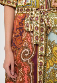 Suurendatud vaade mustrilisele riidekleidile, kus on paisley ja lillemotiivid oranžis, kollases, lillakas ja rohelises toonis, millel on seotud vöö ja lähikonnas resting käsi.