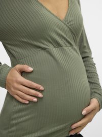 Zelen top z rebrastim vzorcem in dolgimi rokavi, z V-izrezom in nabranim pasom, nošen prek zaobljenega trebuha, roke nežno počivajo na njem.