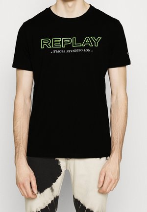 T-shirt noire avec texte "REPLAY" en néon vert et texte blanc à l'envers en dessous, porté avec un pantalon tie-dye noir et blanc.