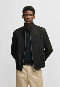 Schwarze Zip-Jacke mit hohem Kragen, elastischen Bündchen und Seitentaschen. Über einem dunklen Oberteil getragen, kombiniert mit hellbeigen Hosen.