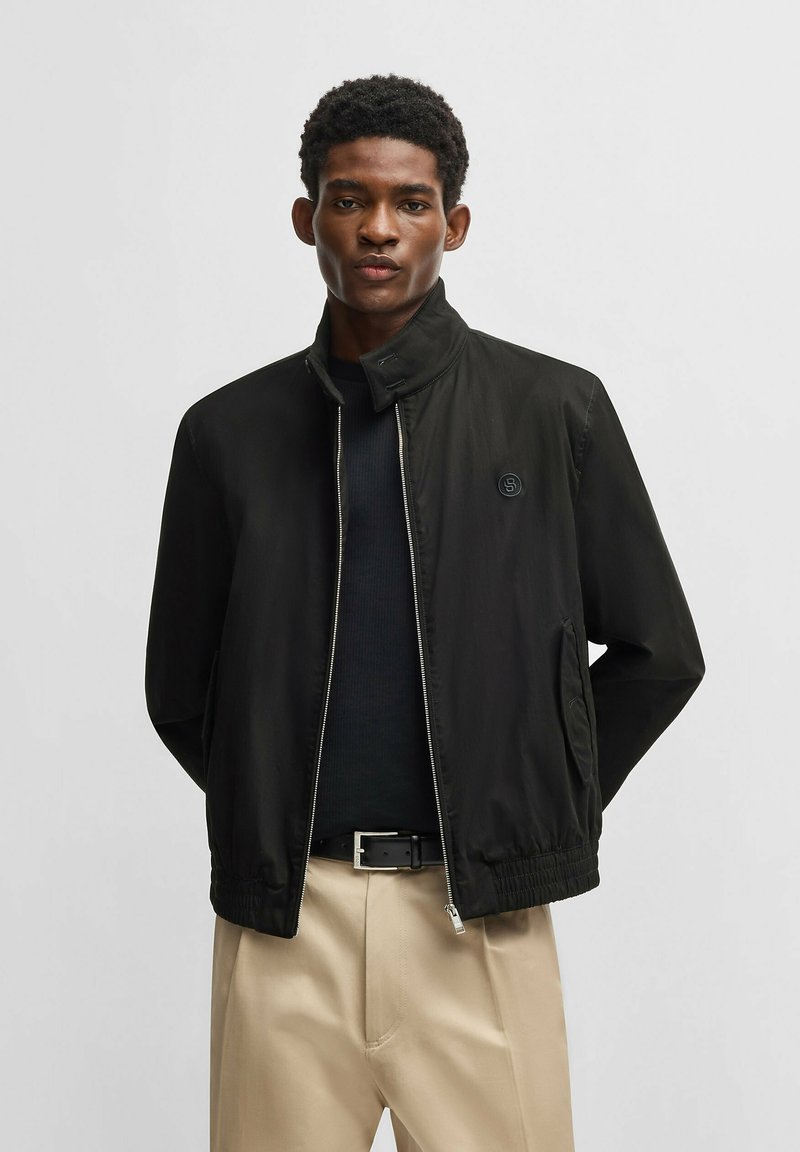 Schwarze Zip-Jacke mit hohem Kragen, elastischen Bündchen und Seitentaschen. Über einem dunklen Oberteil getragen, kombiniert mit hellbeigen Hosen.