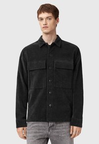 Black corduroy shirt with a shirt collar, two front pockets, and button closure. Features long sleeves and a textured fabric finish.